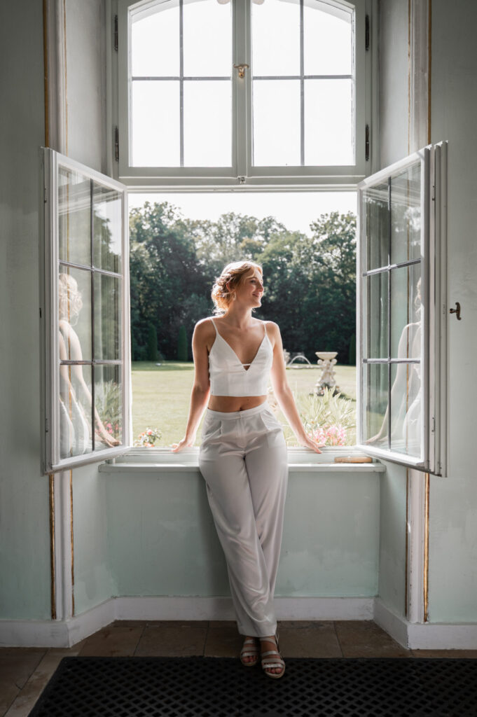 Eine Frau in weißem Outfit steht vor einem offenen Fenster, das Sonnenlicht strömt herein, und draußen liegt ein grüner Garten - die Eleganz der Brautmode aus einem Nürnberger Brautmodenladen.