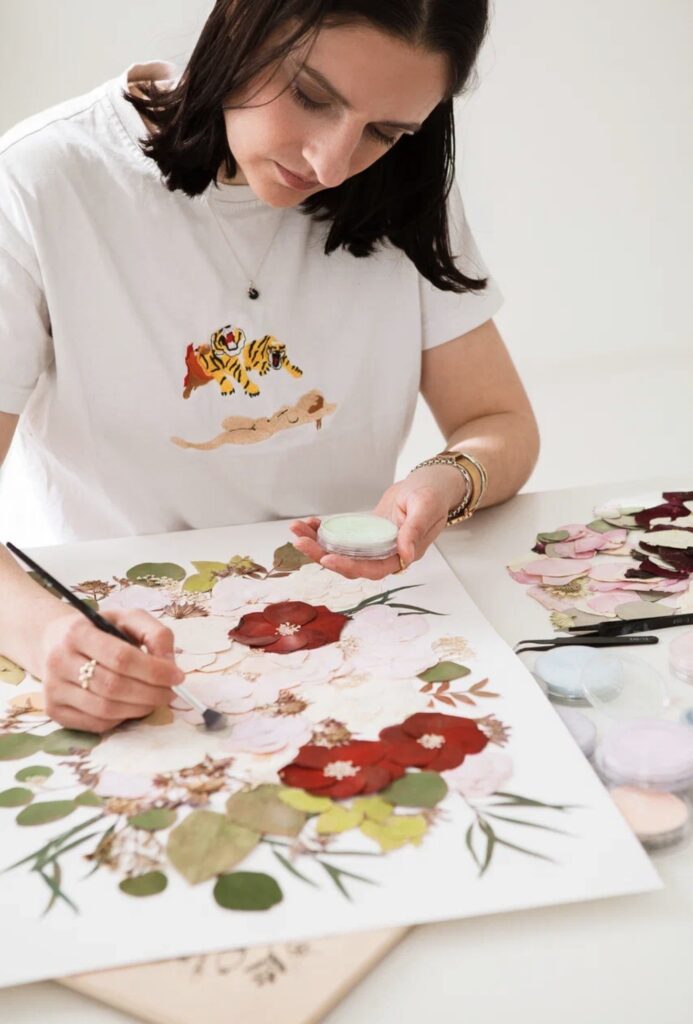 Eine Frau in Nürnberg malt an ihrem Schreibtisch ein florales Kunstwerk, umgeben von Kunstzubehör, gepressten Blumen und Mix & Match-Inspirationen für Brautmode.