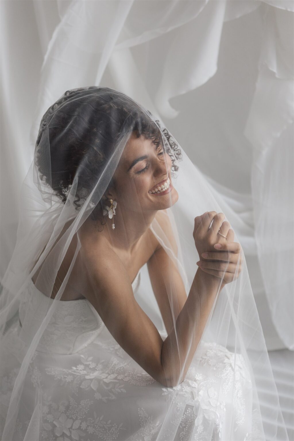 Eine lächelnde Braut in weißem Spitzenkleid und Schleier, sitzend und sich an den Händen haltend, umgeben von weichem weißem Stoff - so sieht die Eleganz des Brautmodenladens Mix & Match in Nürnberg aus.