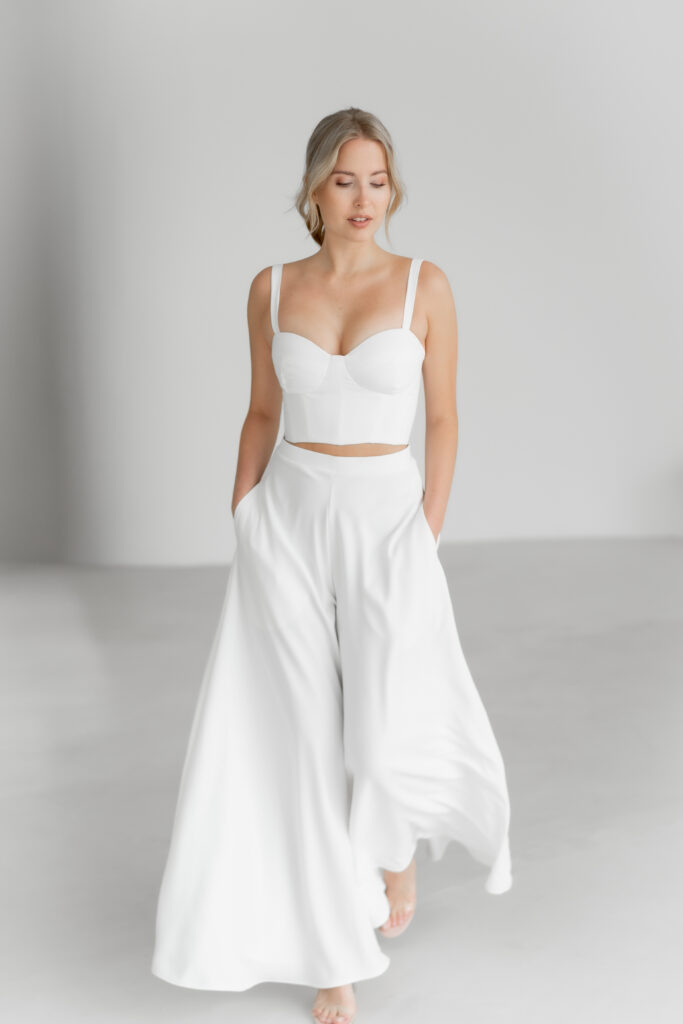 Eine Frau in einem weißen Crop-Top und einer weiten Hose läuft barfuß vor einem hellen, minimalistischen Hintergrund und strahlt mühelose Eleganz aus - die perfekte Inspiration für moderne Brautmode.
