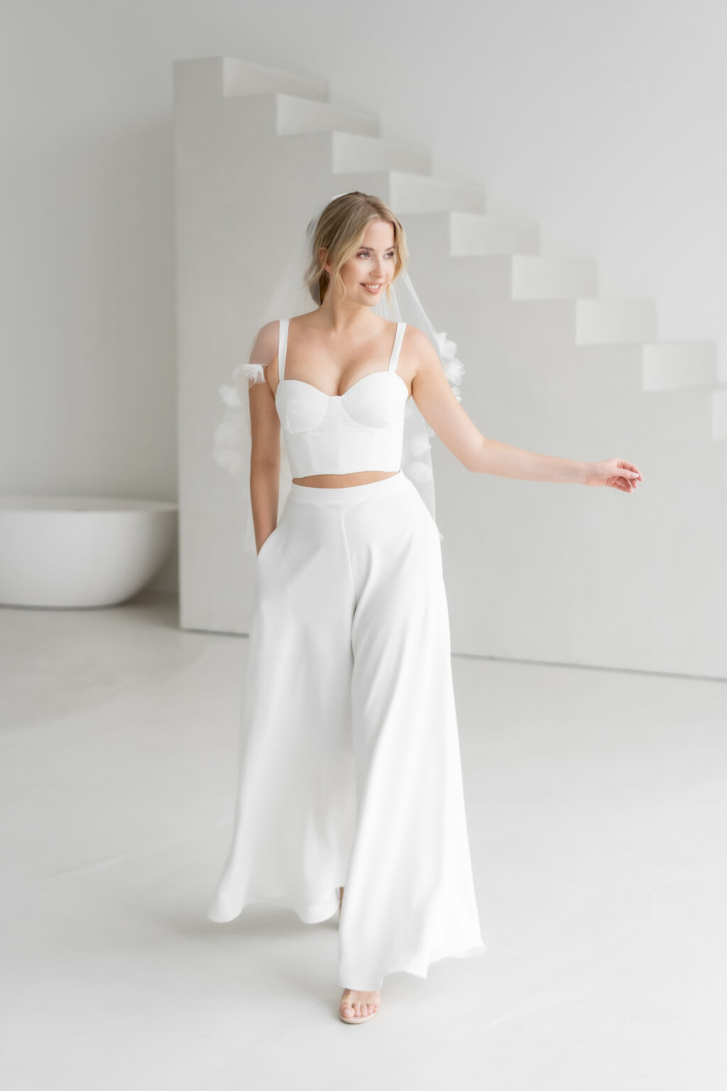 Eine Braut in einem weißen Crop-Top und einer Hose mit weitem Bein posiert in einem minimalistischen, modernen Raum mit weißer Treppe und präsentiert ein Mix & Match Brautoutfit, das ideal für die stilvolle Nürnberger Braut ist.