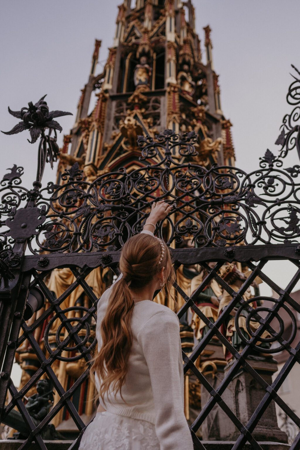 Eine Frau in Weiß berührt einen verschnörkelten schwarzen Zaun und blickt auf einen hohen, dekorativen Turm im gotischen Stil - ihre Brautmode spiegelt elegant einen Mix & Match-Stil wider.