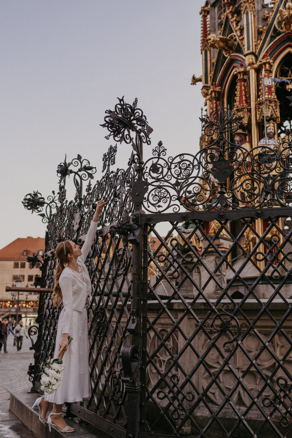 Eine Frau in einem weißen Brautoutfit hält Blumen in die Höhe und streckt sich nach einem verzierten schwarzen Eisenzaun in der Nähe eines historischen Denkmals.