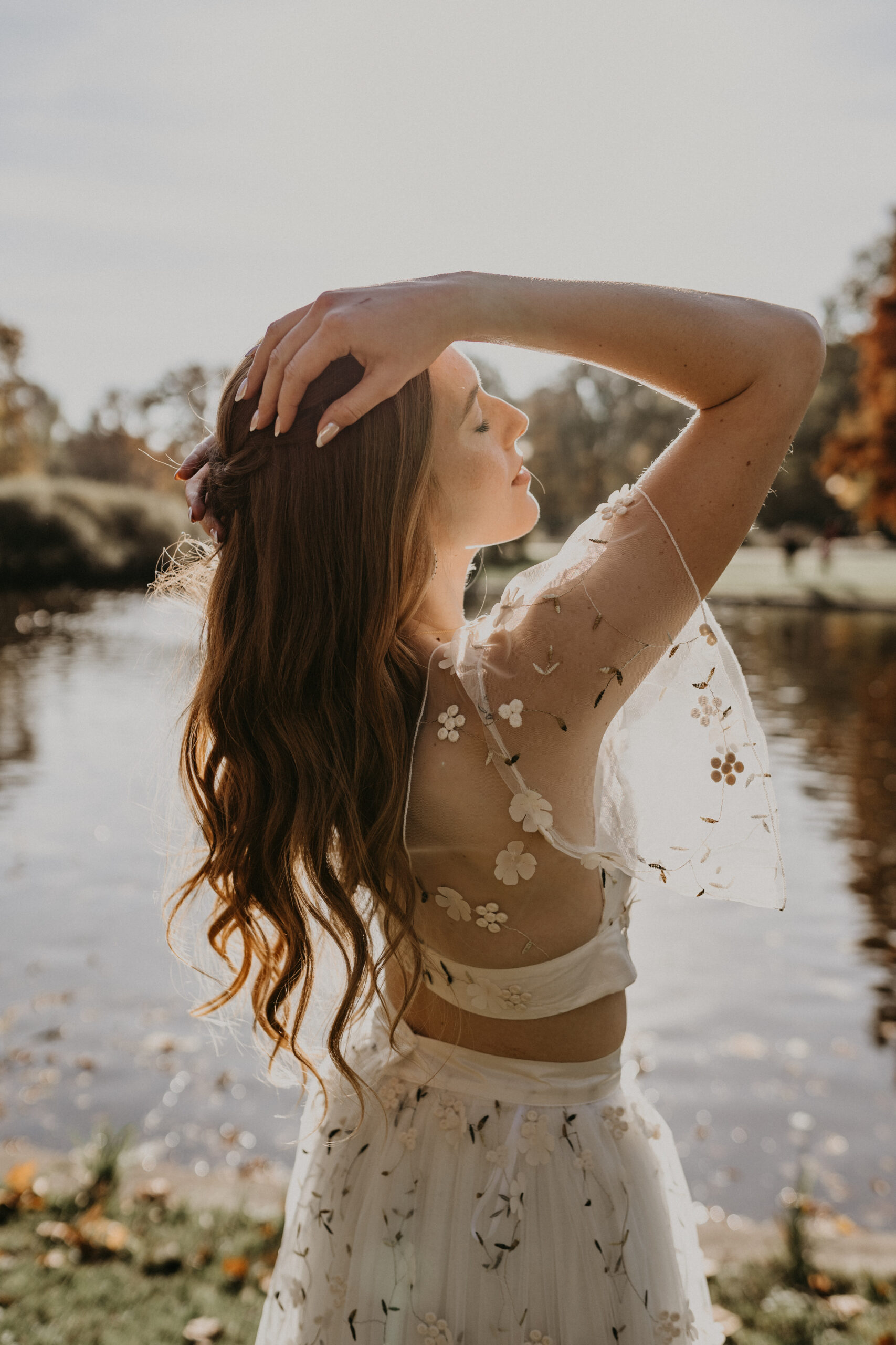 Eine Frau in einem geblümten Kleid posiert an einem sonnenbeschienenen See in Nürnberg, die Augen geschlossen, die Hand auf dem Kopf ruhend - die perfekte Inspiration für Ihr nächstes Mix & Match Brautoutfit.