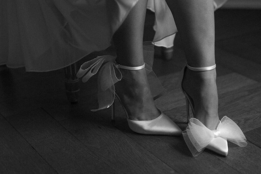 Eine Person im schicken Brautoutfit trägt Satin-Highheels mit großen Tüllschleifen und steht in einem stilvollen Kleid auf einem Holzboden - die perfekte Inspiration für Brautmode-Liebhaber in Nürnberg.