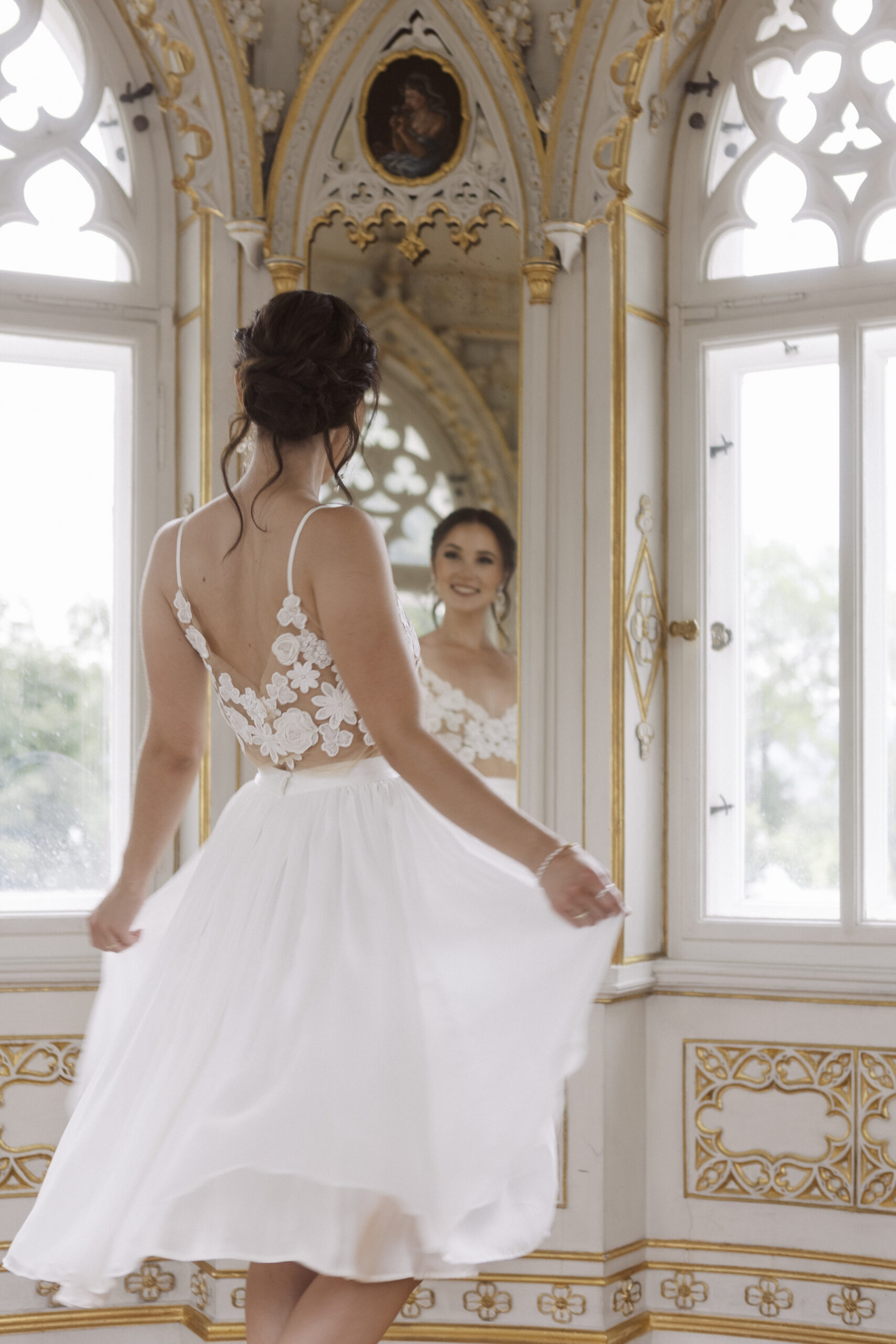 Eine Frau in einem weißen Brautmode-Kleid wirbelt vor einem Spiegel in einem verzierten Raum mit Bogenfenstern und fängt die Eleganz des Nürnberger Mix & Match-Brautstils ein.
