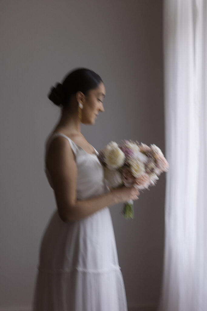 Eine Frau in einem weißen Brautmodenkleid hält einen Blumenstrauß in der Hand, während sie an einem Fenster mit transparenten Vorhängen steht und die Eleganz eines Top-Brautmodenladens in Nürnberg einfängt.