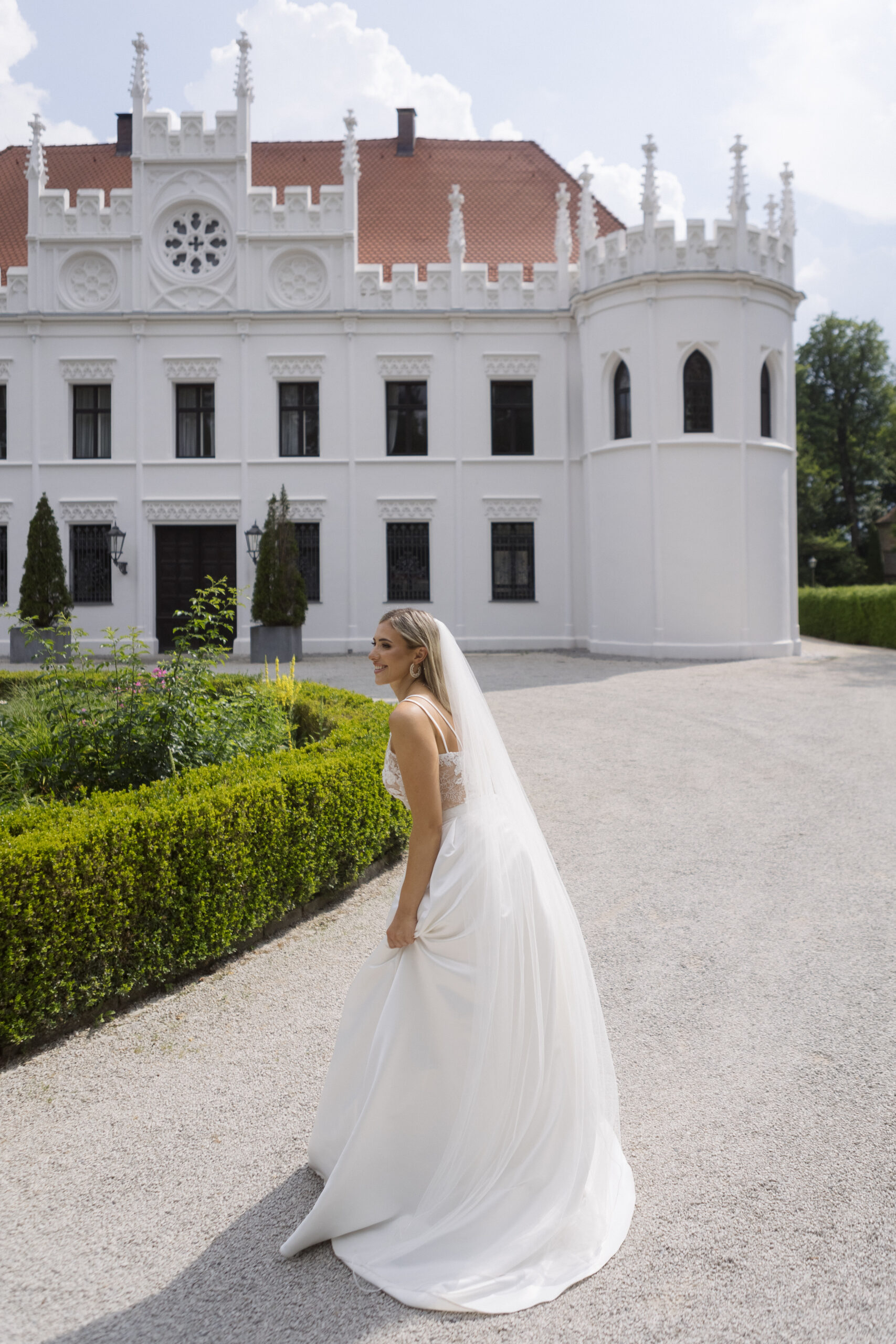 Eine Braut in eleganter Brautmode, mit weißem Kleid und Schleier, steht vor einem großen, weißen historischen Gebäude mit rotem Dach.