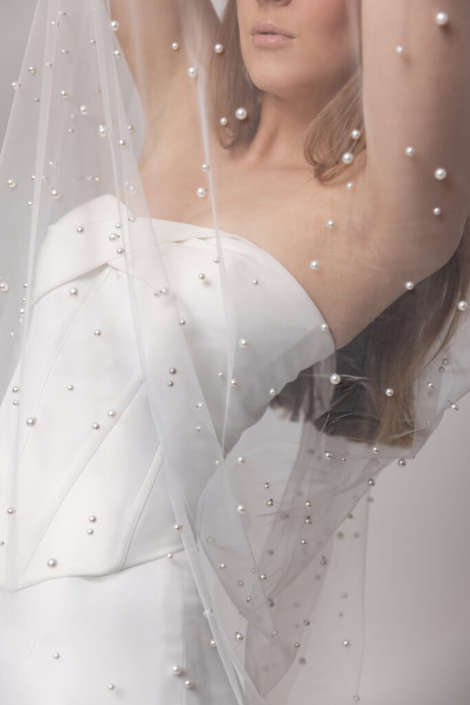 Eine Frau in einem weißen trägerlosen Kleid mit einem durchsichtigen, perlenbesetzten Schleier über dem Oberkörper zeigt elegante Brautmode, perfekt für ein zeitloses Brautoutfit in Nürnberg.