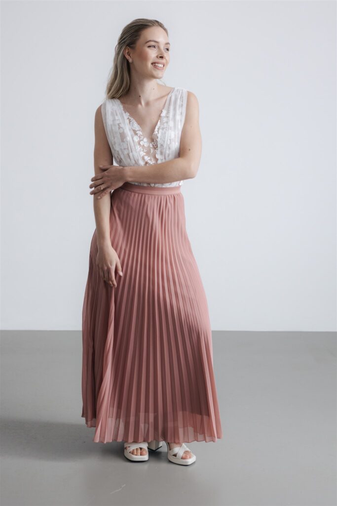 Eine Frau in einem weißen ärmellosen Oberteil und einem langen rosa Faltenrock, stehend und lächelnd - die perfekte Inspiration für ein modernes Brautoutfit in Nürnberg.