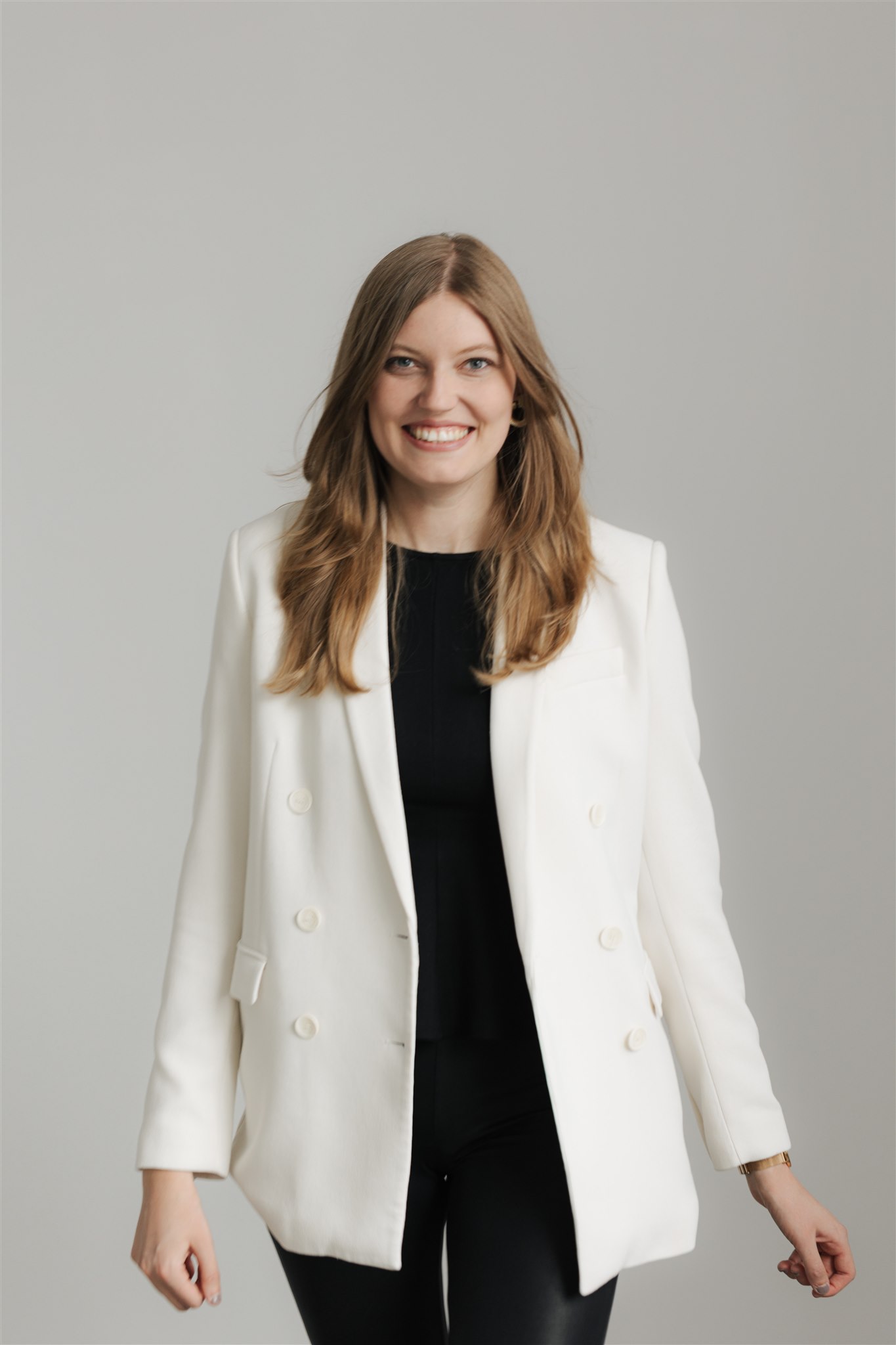 Laura Zeltner in weißem Blazer und schwarzem Outfit, die ein schickes Mix & Match Brautoutfit präsentiert, steht vor einem schlichten hellgrauen Hintergrund.
