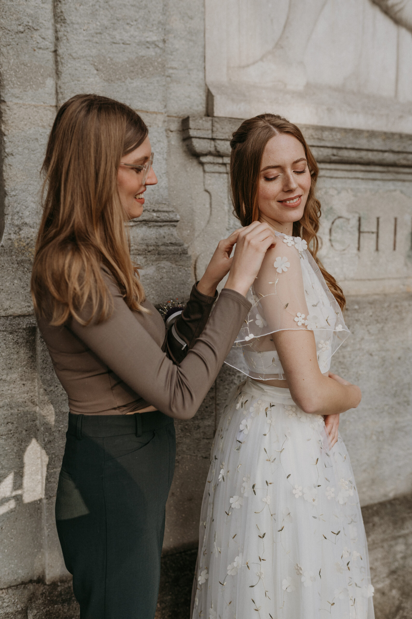 Eine Frau hilft einer anderen Frau, ihr geblümtes Brautmode-Brautkleid im Freien in Nürnberg anzupassen. Beide stehen lächelnd an einer Steinmauer und genießen die Freude am Mix & Match-Stil.