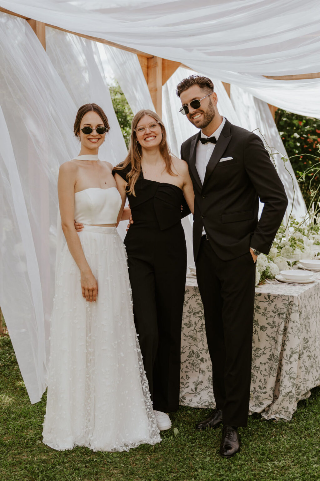 Drei formell gekleidete Personen posieren und lächeln im Freien vor einer mit weißem Stoff drapierten Hochzeitsdekoration, die elegante Brautmode für jede Nürnberger Feier präsentiert.