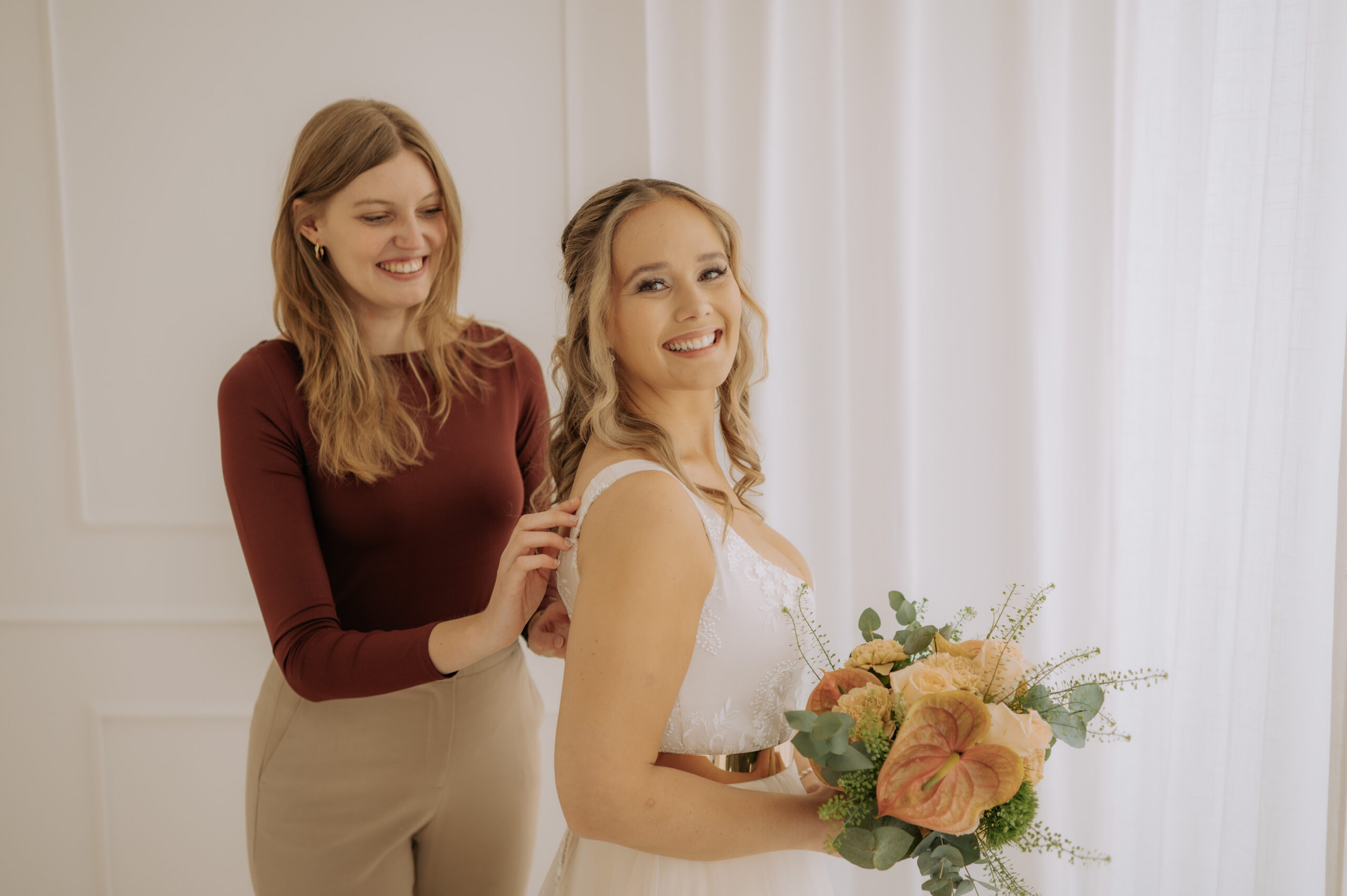 Eine lächelnde Braut mit einem Blumenstrauß in der Hand, während eine andere Frau ihr Brautoutfit in einem hellen Raum zurechtrückt, fängt einen fröhlichen Moment in der Nürnberger Welt der Brautmode ein.