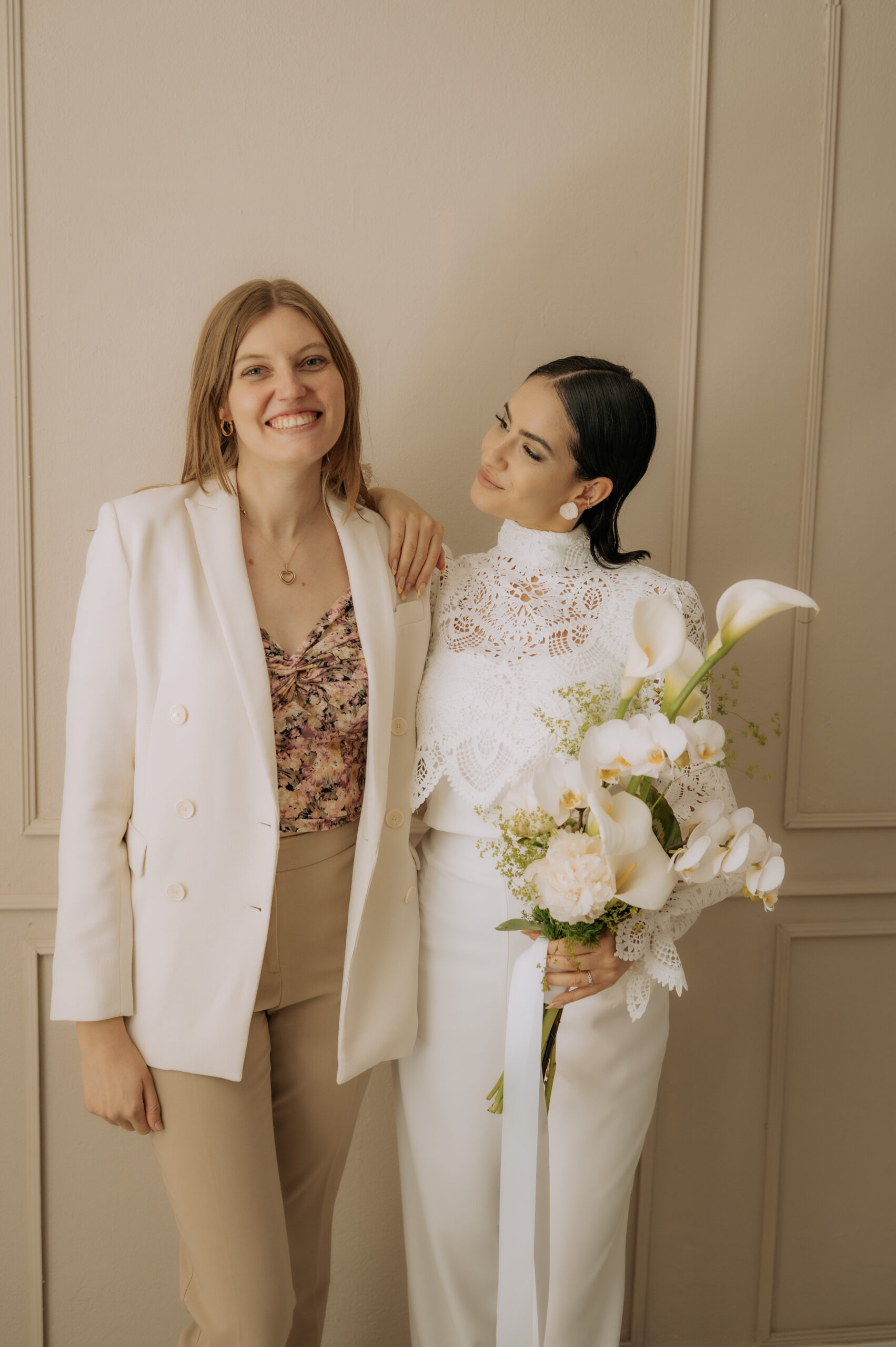 Zwei Frauen posieren zusammen; die eine im weißen Spitzenkleid mit Blumen in der Hand, die andere im beigen Anzug und geblümtem Oberteil - ein Mix & Match-Stil, perfekt für jeden Brautmodenladen in Nürnberg.