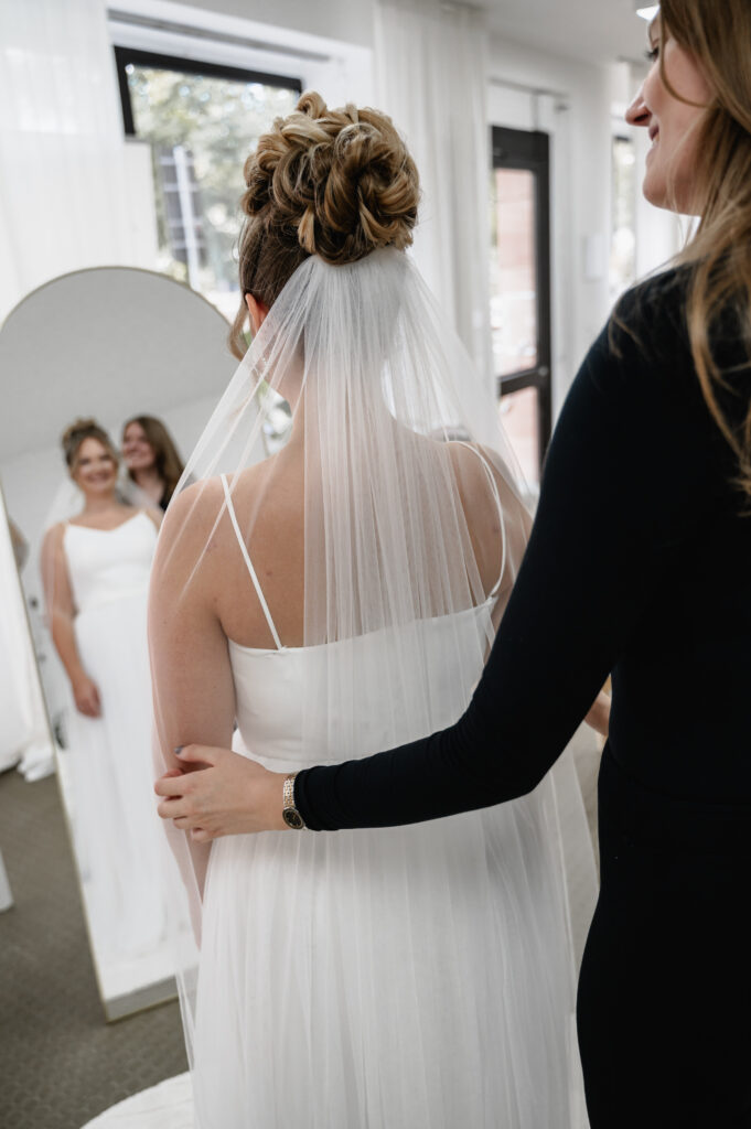 Eine Braut in weißem Kleid und Schleier steht mit einer Frau in einem Brautmodenladen in Nürnberg und bewundert ihr Spiegelbild, während sie ihr Brautoutfit perfektioniert.