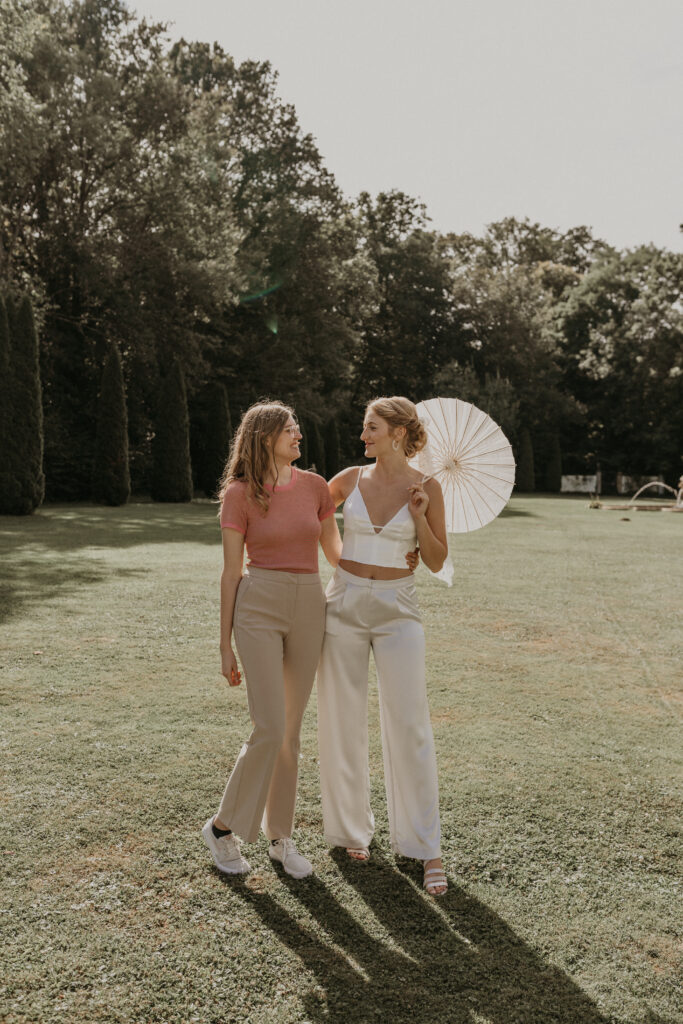 Zwei Frauen, die sich an einem sonnigen Tag auf einer Wiese in Nürnberg anlächeln; eine hält einen Sonnenschirm aus Papier in der Hand. Ihre Mix & Match-Outfits sorgen für einen frischen Look, perfekt, um gemeinsam in einem Brautmodenladen zu stöbern.