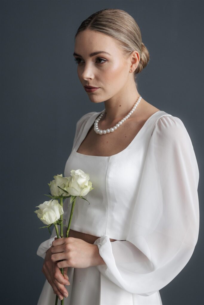Frau im weißen Kleid, weiße Rosen haltend, mit Perlenkette, die Haare zu einem tiefen Dutt gebunden - die perfekte Inspiration für Ihr Brautoutfit aus dem Brautmodenladen.