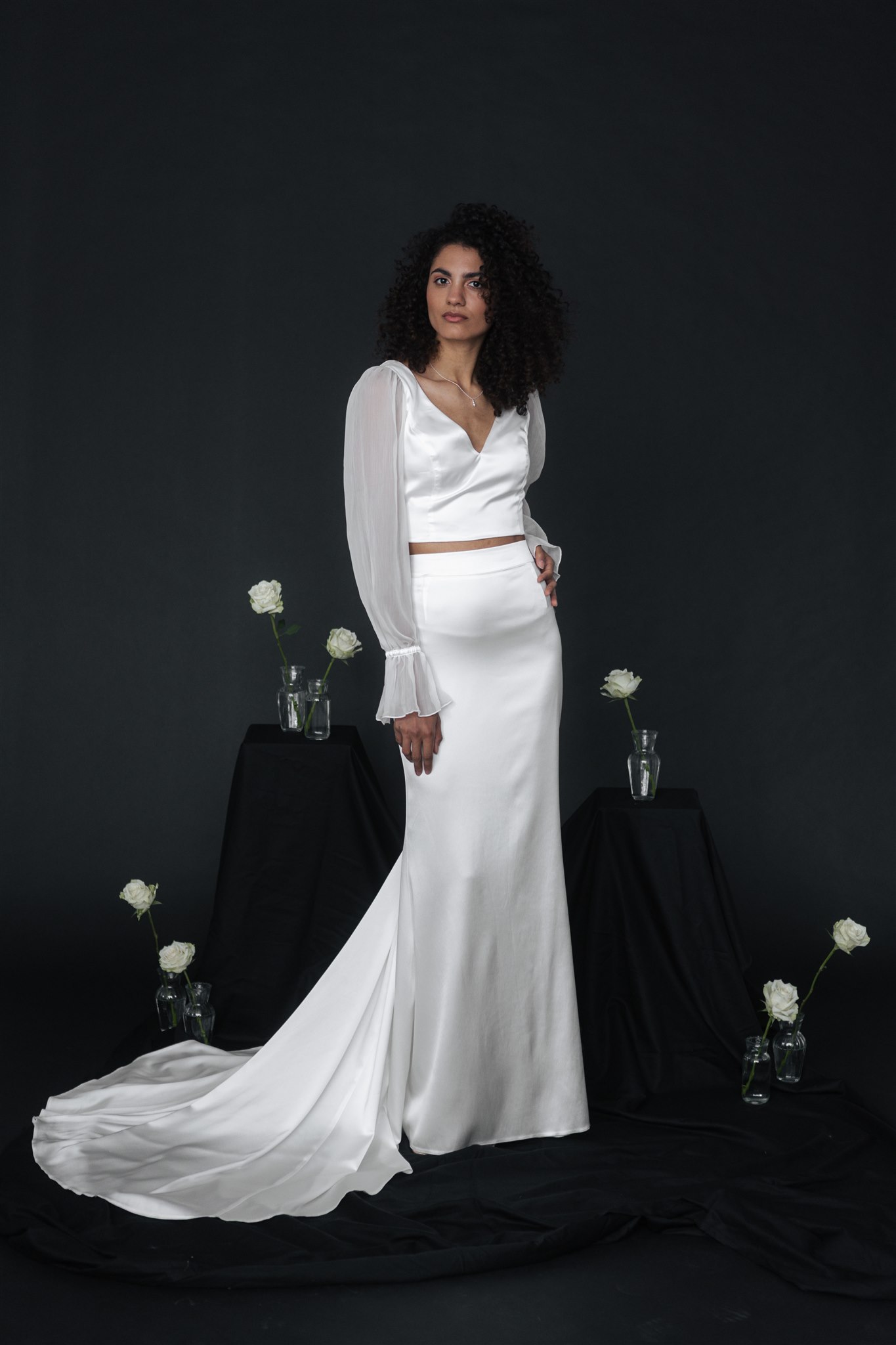 Eine Frau im weißen Satin-Brautoutfit steht zwischen Glasvasen mit weißen Blumen vor einem dunklen Hintergrund - die perfekte Inspiration für elegante Brautmode in Nürnberg.