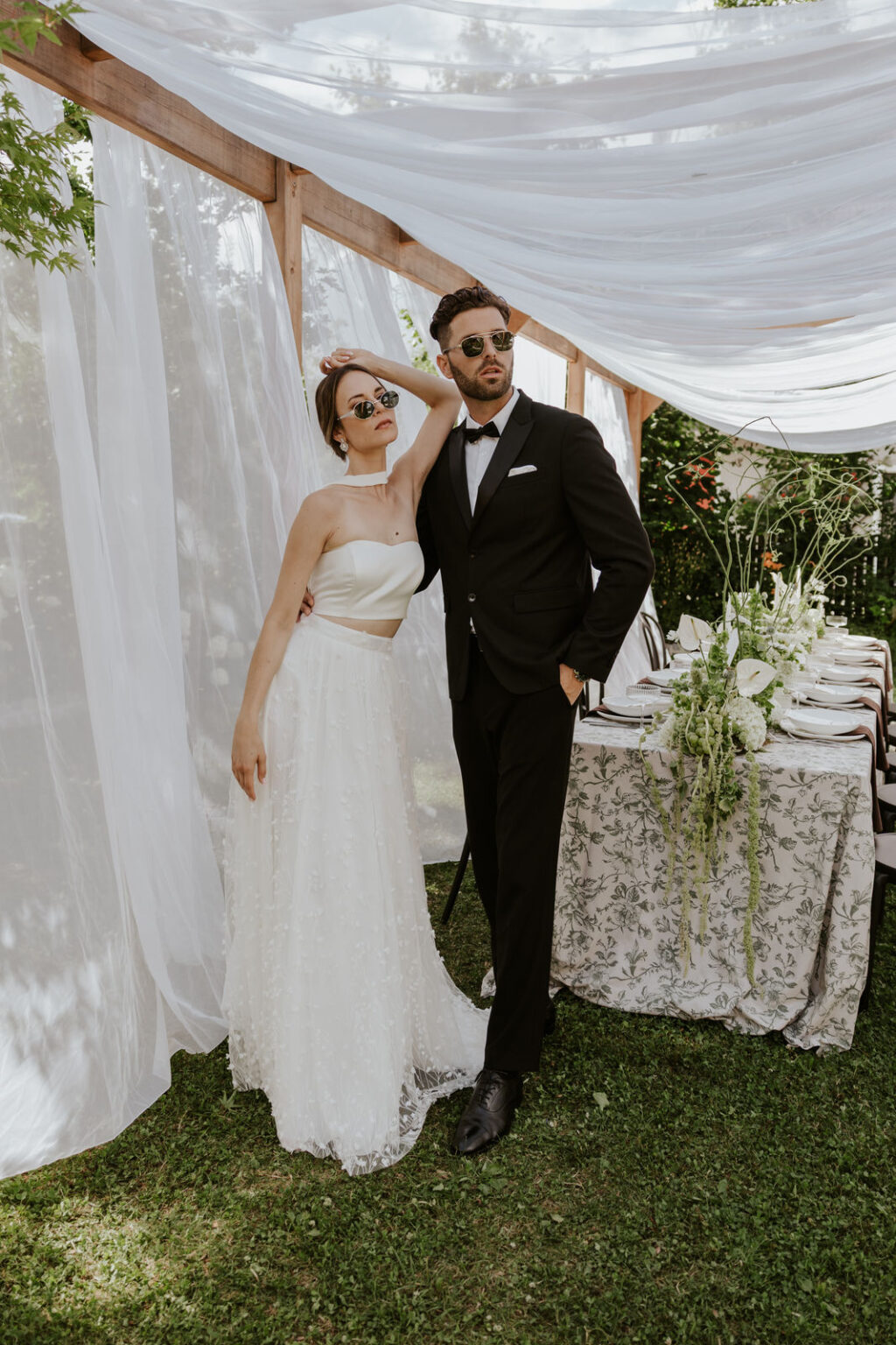 Braut und Bräutigam mit Sonnenbrille posieren stilvoll unter einem weißen Baldachin neben einem eleganten, blumengeschmückten Tisch - und fangen damit moderne Brautmode-Vibes ein, die perfekt zur Inspiration für jeden Nürnberger Brautmodenladen sind.