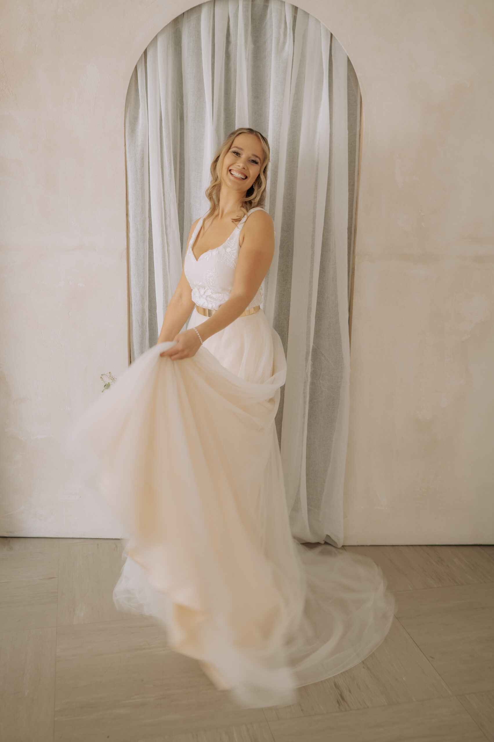 Eine lächelnde Frau in einem weißen Hochzeitskleid wirbelt vor einem durchsichtigen Vorhang und einer gewölbten Tür und präsentiert elegante Brautmode in Nürnberg im schicken Mix & Match-Stil.