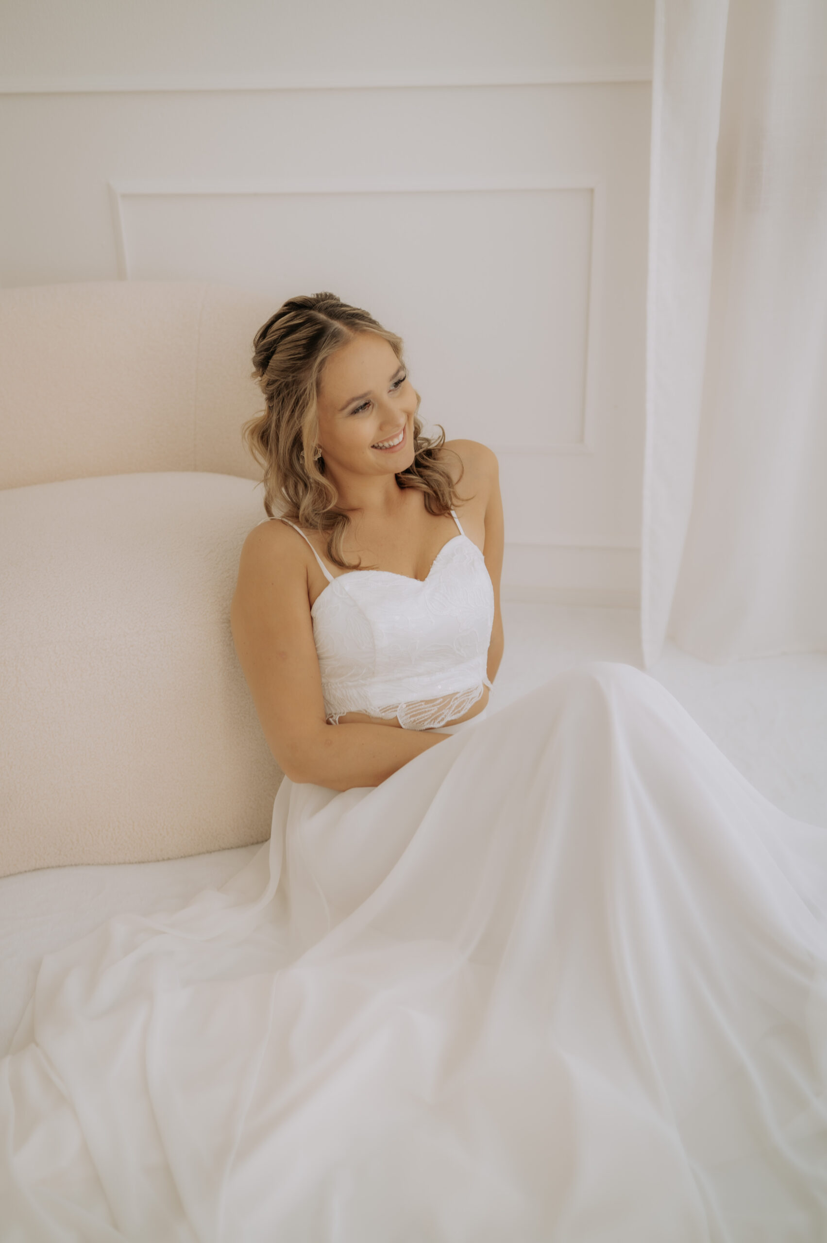Eine lächelnde Frau in einem weißen Brautkleid aus einer Mix & Match Brautmode-Kollektion sitzt auf dem Boden und lehnt sich an ein helles Sofa in Nürnberg.
