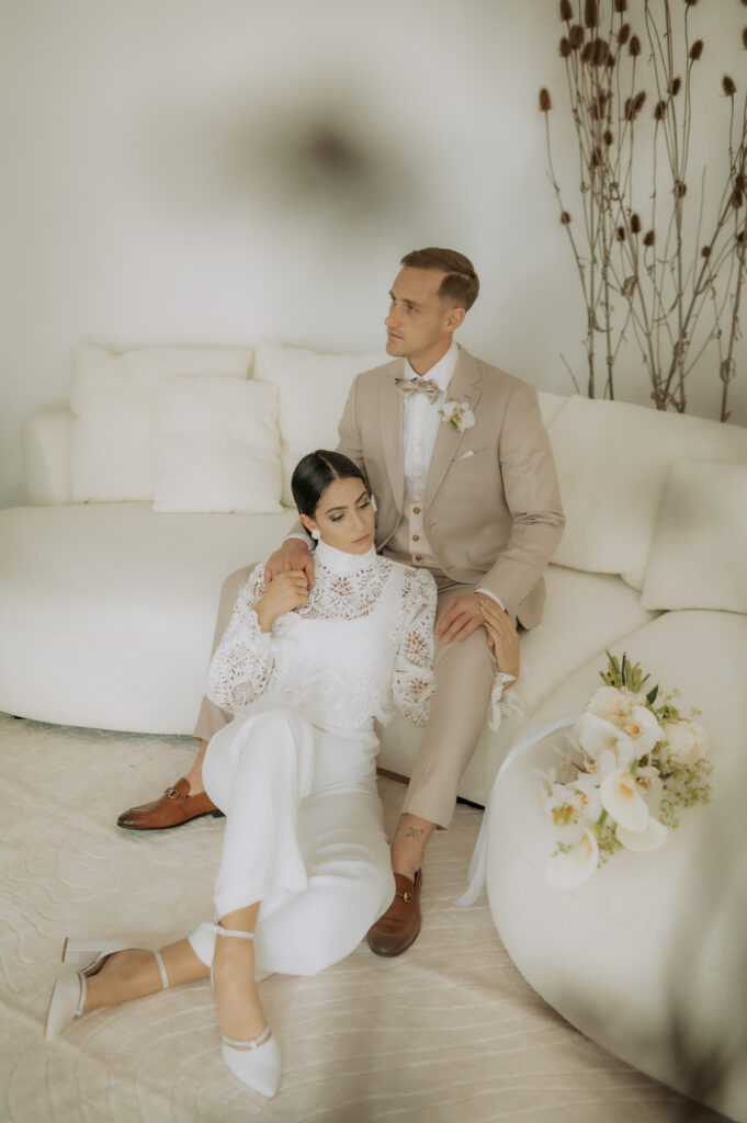 Eine Braut in Weiß zeigt elegante Brautmode, während sie auf dem Boden sitzt, angelehnt an einen Bräutigam in einem beigen Anzug, mit einem Blumenstrauß in der Nähe auf einem weißen Sofa - und fängt damit zeitlose Liebe in Nürnberg ein.
