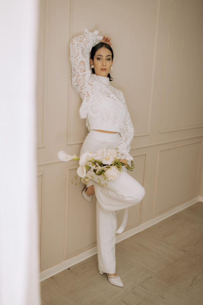 Eine Frau in weißem Spitzentop und Hose posiert in einem Innenraum mit einem Blumenstrauß vor einer beigen Wand und präsentiert damit moderne Brautmode und Mix & Match-Stil in Nürnberg.