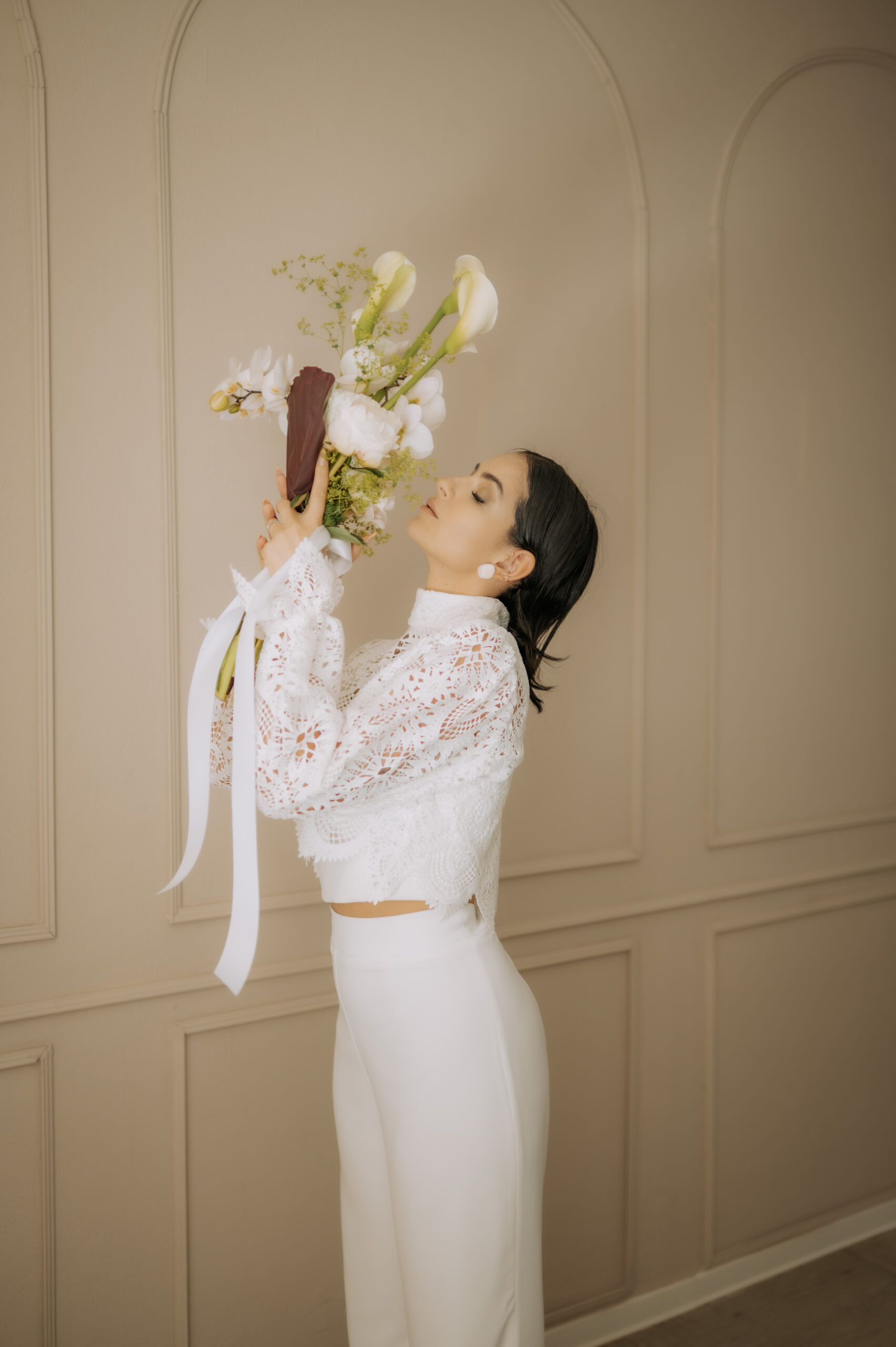 Frau in weißem Spitzen-Brautoutfit und Hose hält einen Blumenstrauß und steht seitlich vor einer beigen Wand.