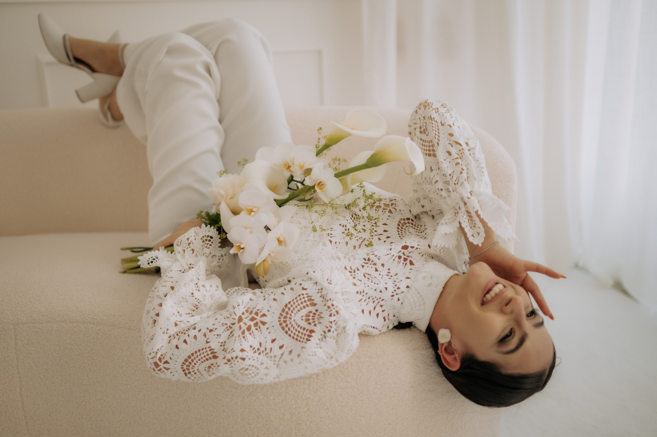 Frau in weißem Spitzenoutfit liegt auf einem cremefarbenen Sofa, hält weiße Blumen in der Hand, lächelt und sieht entspannt aus - die perfekte Inspiration für Mix & Match Styles in jedem Brautmode oder Brautmodenladen.