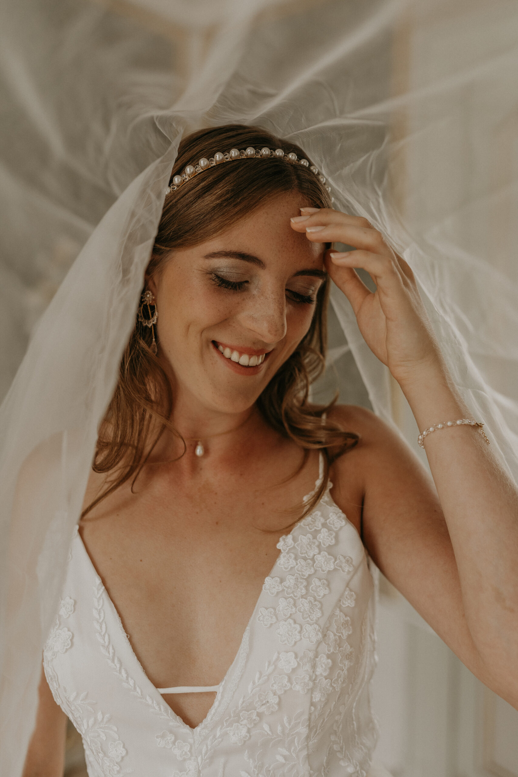 Lächelnde Braut in weißem Kleid und Schleier, die ihr Haar mit einem Perlenstirnband und Schmuck berührt - das perfekte Mix & Match Brautoutfit für elegante Brautmode.