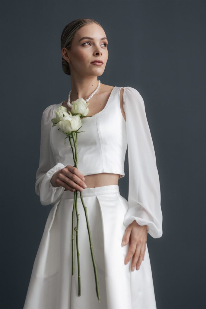 Eine Frau in einem weißen Kleid mit weißen Rosen vor einem dunkelgrauen Hintergrund - die perfekte Inspiration für Mix & Match Styles in Ihrem Nürnberger Brautmodenladen.