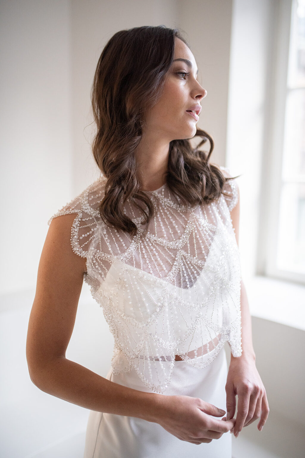 Frau in weißem, perlenbesetztem, ärmellosen Brautkleid aus Nürnberg steht am Fenster und blickt nachdenklich zur Seite - die perfekte Inspiration für elegante Brautmode und Ihr Traum-Brautoutfit.