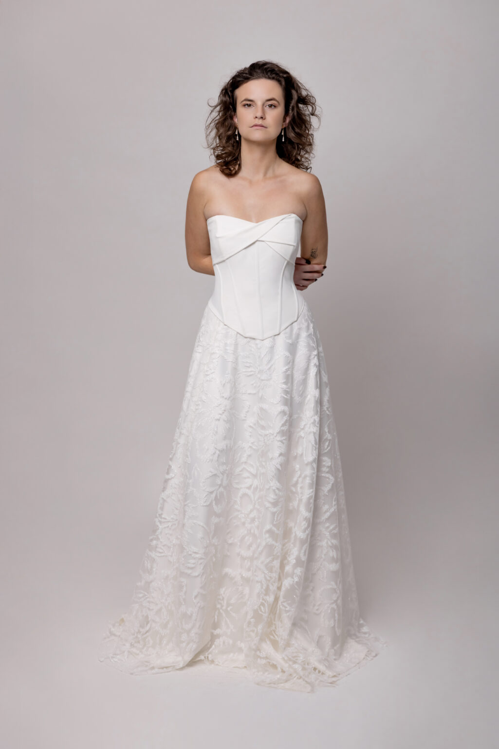 Frau in einem trägerlosen weißen Hochzeitskleid mit tailliertem Mieder und fließendem Spitzenrock, das ein elegantes Brautoutfit aus einem Nürnberger Brautmodenladen präsentiert, vor einem schlichten Hintergrund.