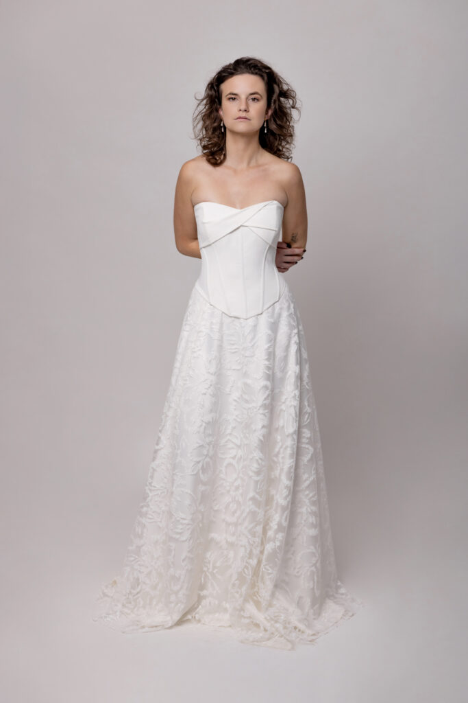 Frau in einem trägerlosen weißen Hochzeitskleid mit tailliertem Mieder und fließendem Spitzenrock, das ein elegantes Brautoutfit aus einem Nürnberger Brautmodenladen präsentiert, vor einem schlichten Hintergrund.