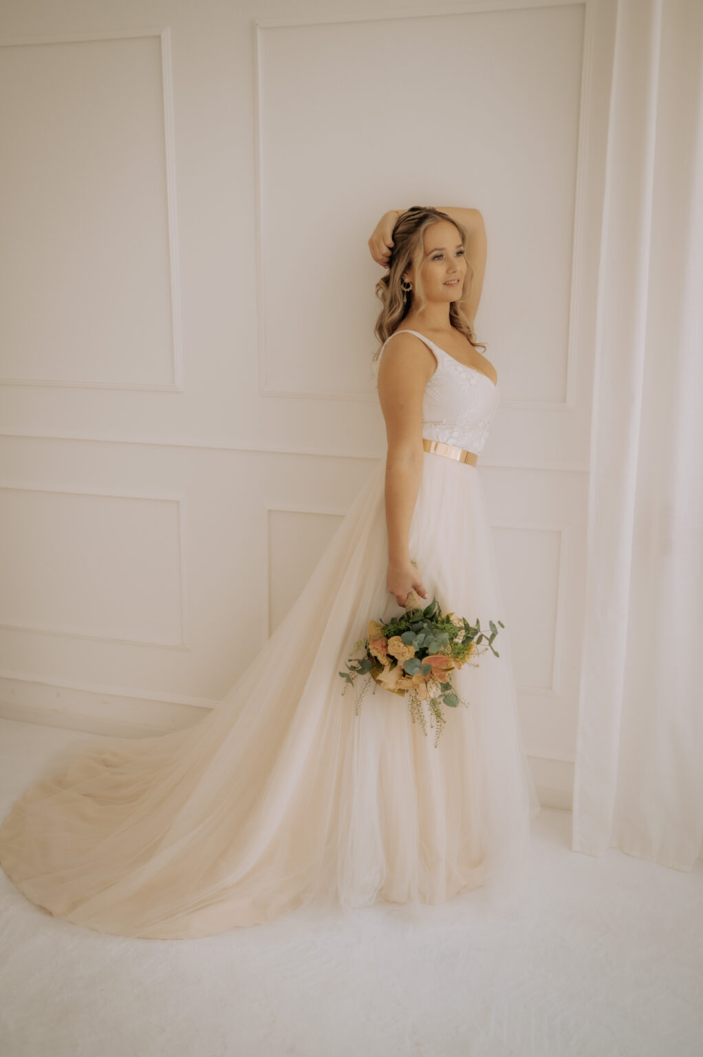 Frau im weißen Brautmode-Brautkleid mit Blumenstrauß vor einer weißen Wand mit sanfter Beleuchtung - perfekt für Bräute in Nürnberg, die den Mix & Match-Stil lieben.