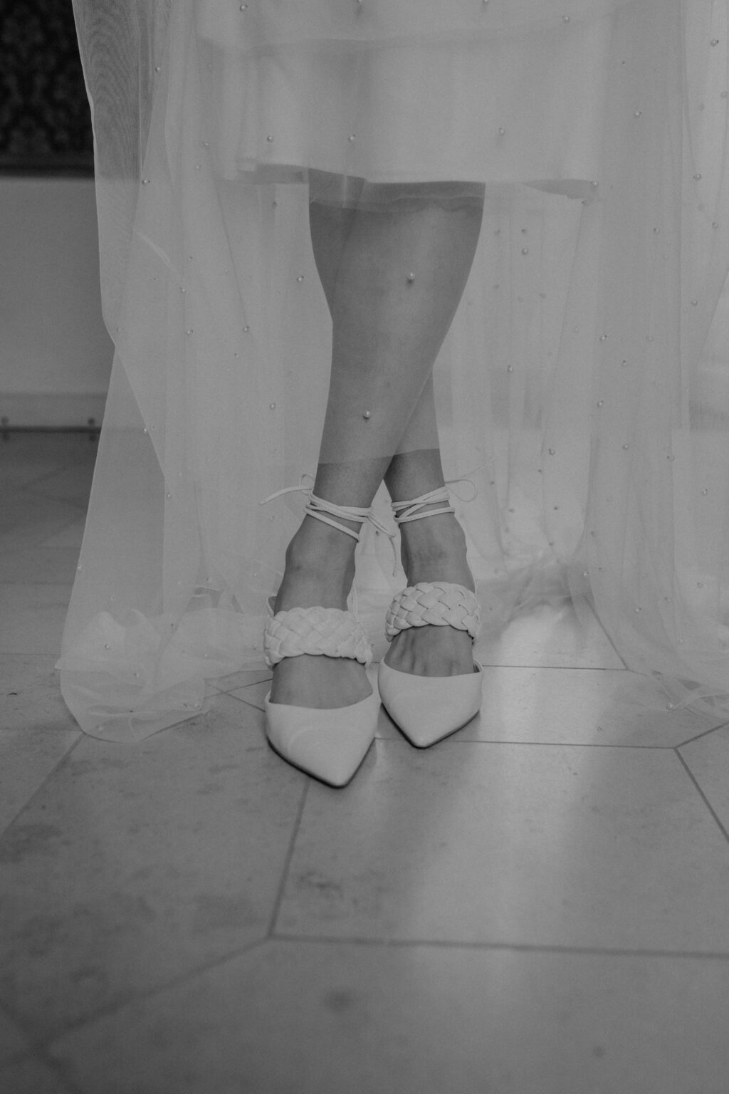 Eine Frau in einem durchsichtigen, perlenbesetzten Brautmodenkleid steht mit gekreuzten Beinen da und trägt spitze, geflochtene Riemenschuhe - eine perfekte Inspiration für einen Nürnberger Brautmodenladen.