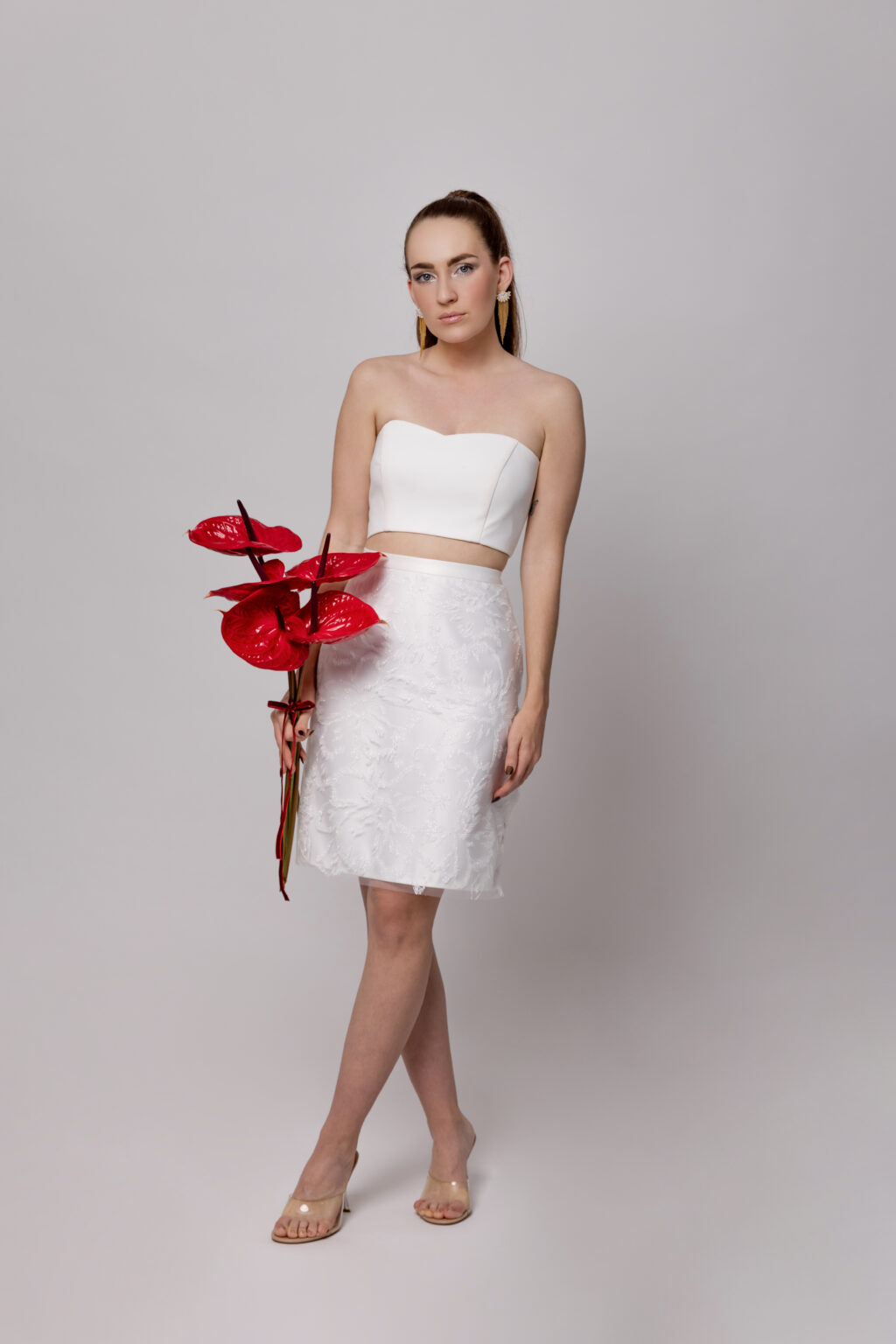 Frau in einem weißen trägerlosen Mix & Match Brautoutfit und Rock, rote Blumen haltend, vor einem schlichten hellen Hintergrund - die perfekte Inspiration für moderne Brautmode.