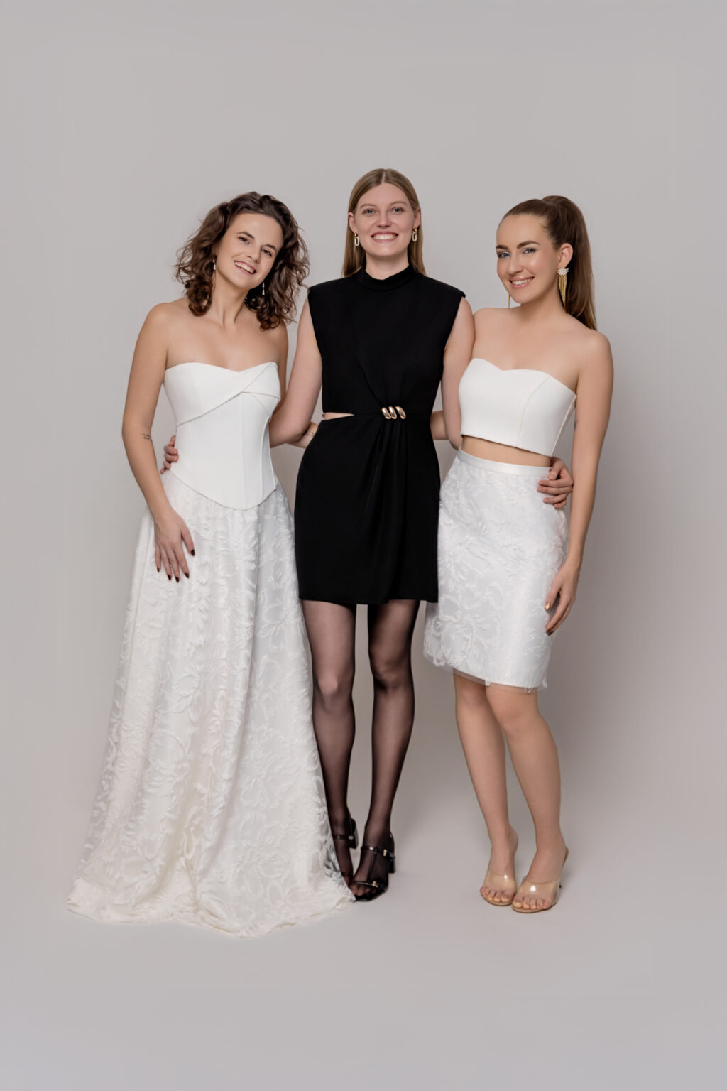 Drei Frauen posieren zusammen, zwei in eleganten weißen Outfits, die an die Brautmode erinnern, und eine in einem ärmellosen schwarzen Kleid mit durchsichtiger schwarzer Strumpfhose - aufgenommen in einem stilvollen Brautmodenladen in Nürnberg.