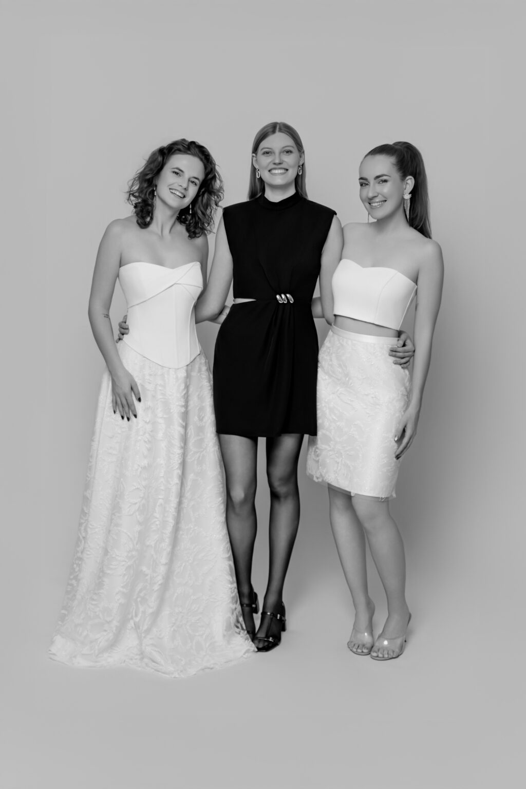 Drei Frauen posieren zusammen in einer Studioumgebung, zwei in weißen Outfits und eine in einem schwarzen Kleid, alle lächelnd - die perfekte Mix & Match Inspiration für jeden Brautmodenladen in Nürnberg.