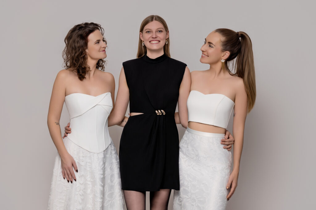 Drei Frauen posieren zusammen; zwei in weißen trägerlosen Brautoutfit-Kleidern, eine in einem schwarzen Kleid, alle lächeln.