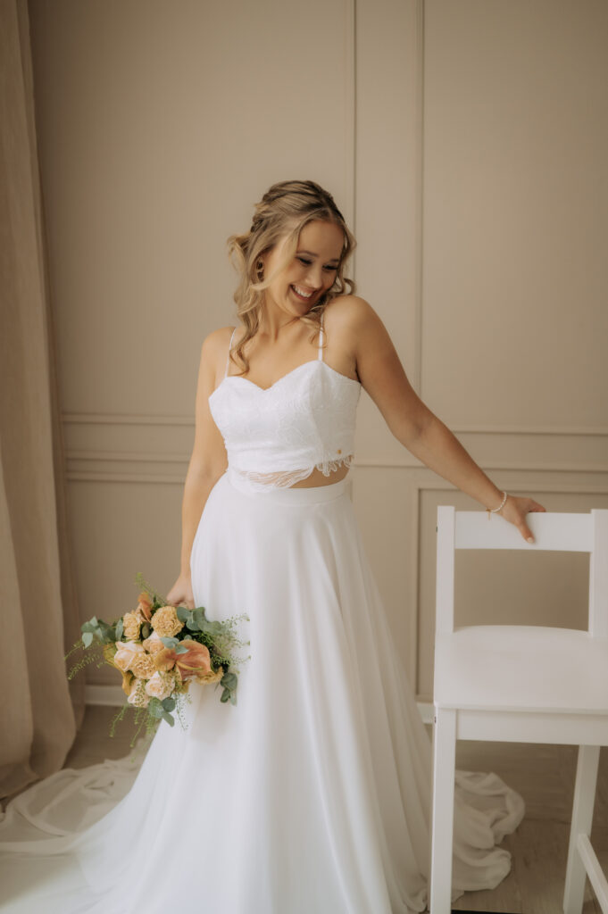 Eine lächelnde Braut in einem umwerfenden Brautoutfit steht neben einem weißen Stuhl in einem sanft beleuchteten Raum und verkörpert den eleganten Mix & Match-Stil, der perfekt für jede Nürnberger Hochzeit ist.