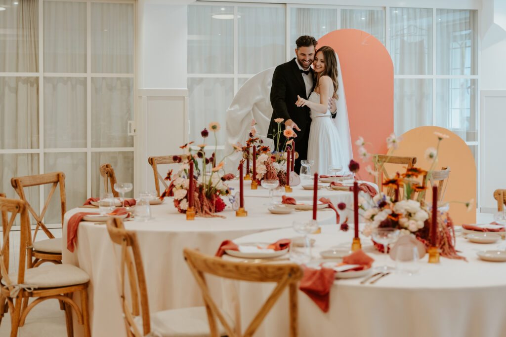 Ein Brautpaar lächelt und umarmt sich hinter einem geschmückten Hochzeitstisch mit Blumengestecken, die die Freude ihrer Hochzeit widerspiegeln und ihr elegantes Brautoutfit zur Geltung bringen.
