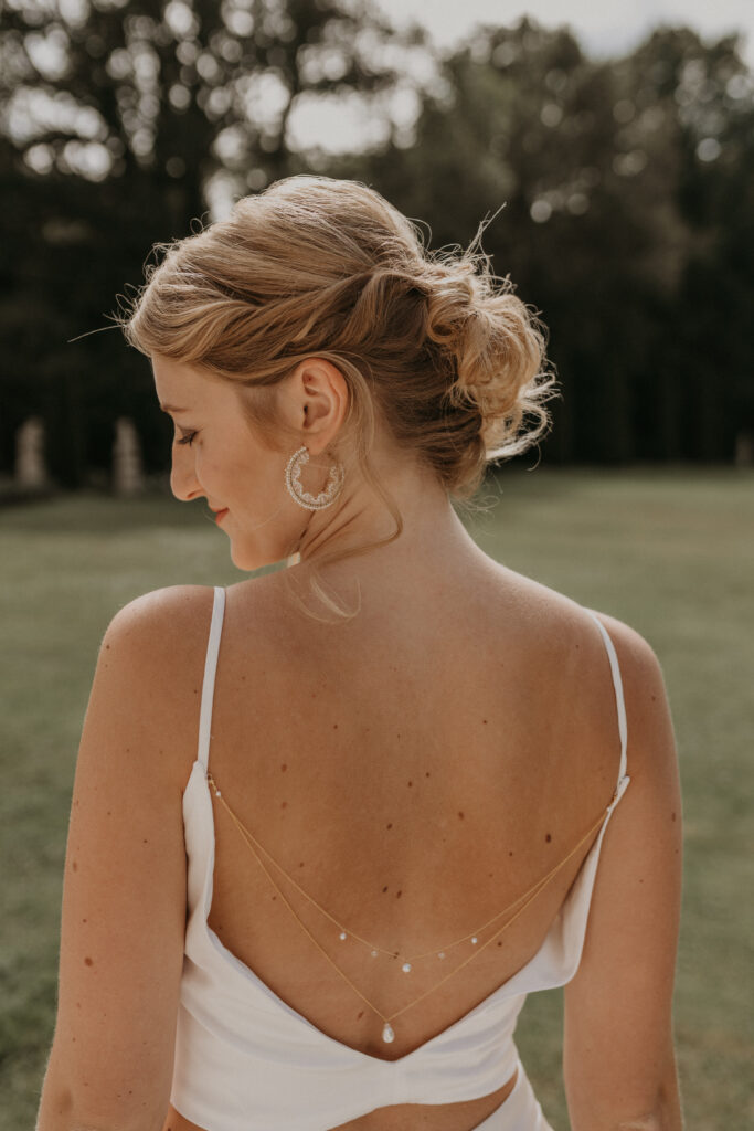Eine Frau mit gestyltem blondem Haar, die ein rückenfreies weißes Kleid und zarten Brautschmuck trägt und im Freien steht.