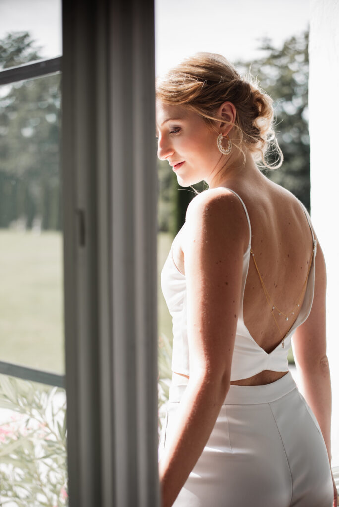 Eine Frau in einem weißen, ärmellosen Kleid steht am Fenster; das Sonnenlicht lässt ihren Brautlook und ihre zarten Accessoires strahlen, während sie nach draußen blickt.