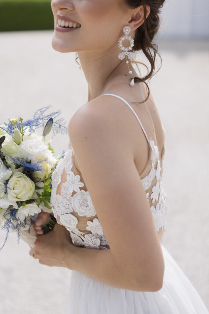 Eine lächelnde Braut in einem Kleid aus Blumenspitze, die einen Strauß aus weißen und blauen Blumen in der Hand hält und auffällige Ohrringe trägt, die ihren Brautlook mit eleganten Accessoires perfekt abrunden.