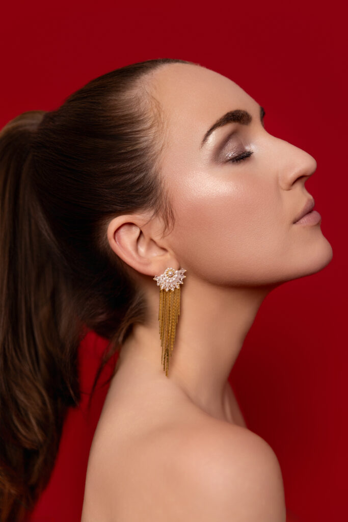 Eine Frau mit Pferdeschwanz und goldenen Ohrringen, geschmückt mit elegantem Brautschmuck, posiert im Profil vor einem leuchtend roten Hintergrund.
