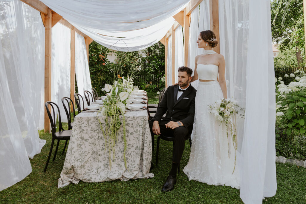 Braut und Bräutigam posieren vor einem elegant dekorierten Hochzeitstisch im Freien und präsentieren unter einem durchscheinenden weißen Baldachin ein stilvolles Brautoutfit und ein romantisches Hochzeitskonzept.