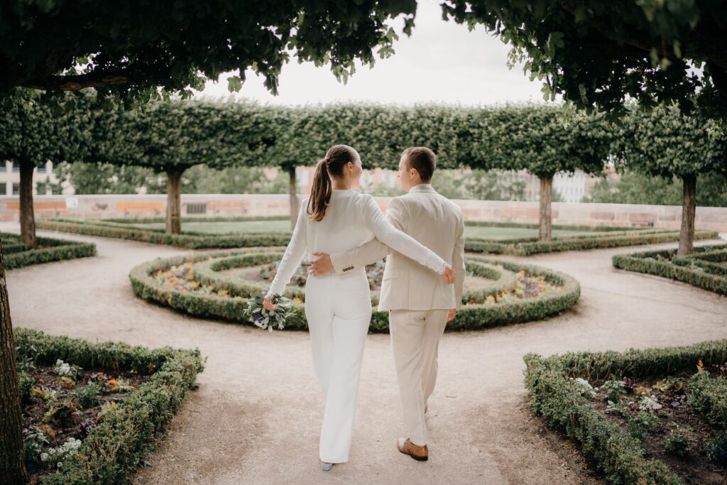 Ein Paar in weißen Outfits spaziert Arm in Arm durch einen gepflegten Garten mit Spiralhecken und zeigt elegante Brautoutfit-Inspirationen, die für einen Nürnberger Brautmodenladen geeignet sind.