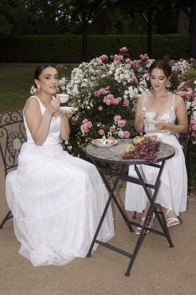 Zwei Frauen in weißen Brautoutfits genießen Tee an einem Gartentisch mit Weintrauben und Gebäck, umgeben von blühenden Rosen – ein traumhaftes Hochzeitskonzept.